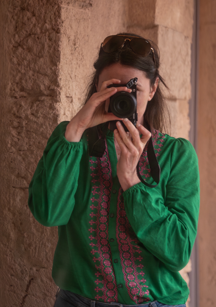 Das Bild zeigt Sandra Biehler. Sie hält eine Kamera vor ihr Gesicht und fotografiert sich selbst im Spiegel. Sie trägt eine grüne Bluse und steht vor einer Naturwand